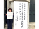 大阪市中央公会堂をバックに表彰状を持つ仲村 千絵代表