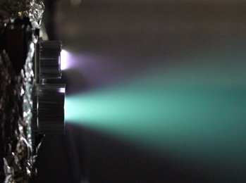 作動中のイオンスラスタを横から撮影した様子。発光部下側はイオンスラスタから発せられるイオンビーム（イオンジェット）、発光部上側は中和器からの電子放出に伴うプラズマ発光（東京大学の発表資料より）