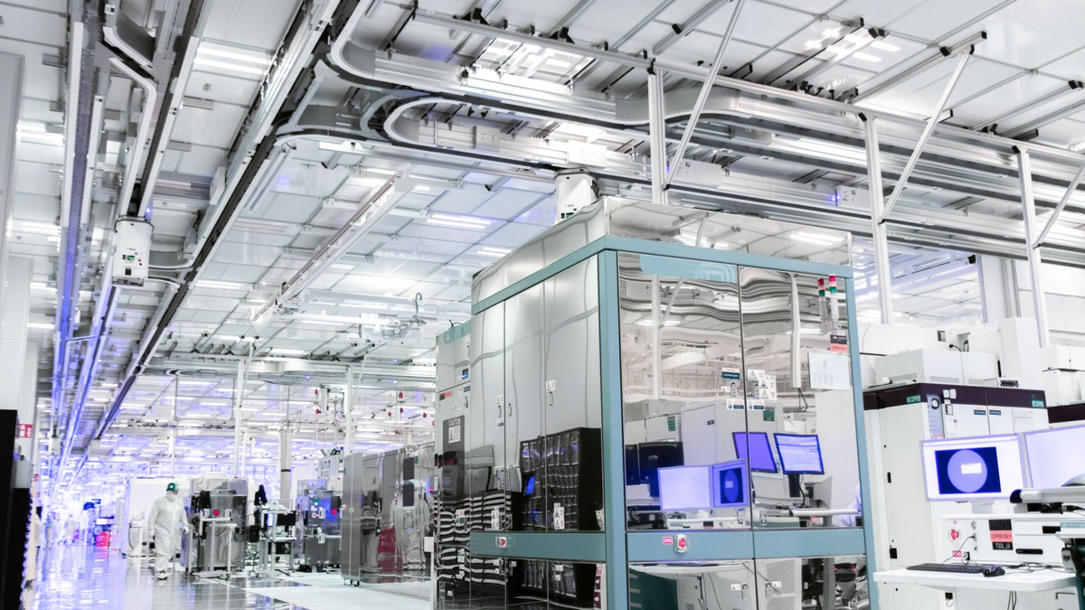 A modern industrial facility interior with advanced machinery and bright lighting. The equipment includes various computer monitors and high-tech apparatus, surrounded by clean, organized workspaces. People in protective suits work in the background. (Intel.com) A modern industrial facility interior with advanced machinery and bright lighting. The equipment includes various computer monitors and high-tech apparatus, surrounded by clean, organized workspaces. People in protective suits work in the background. (Intel.com)